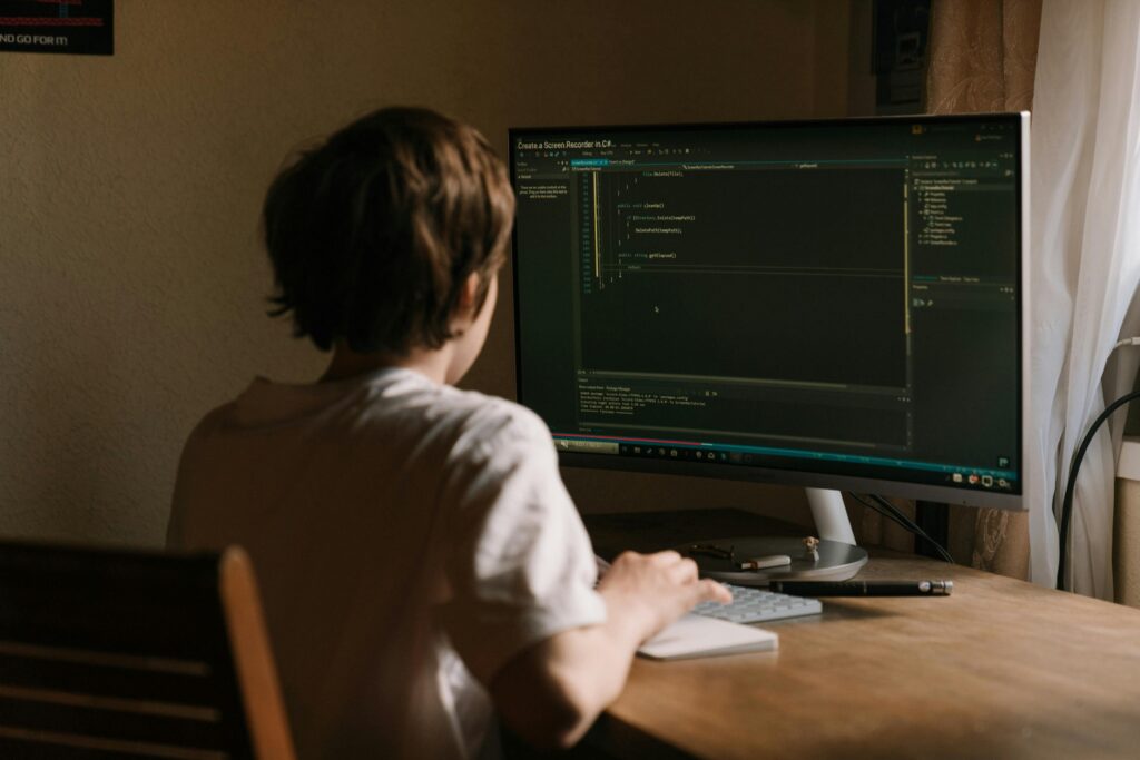 A teenager focused on coding software on a desktop monitor in a home office setting.